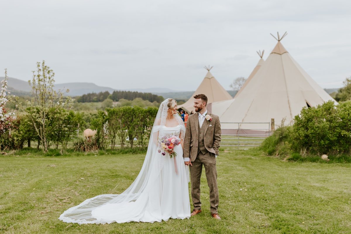 A bride and groom hand in hand on the lawn, with three Java tipis and rolling hills behind them