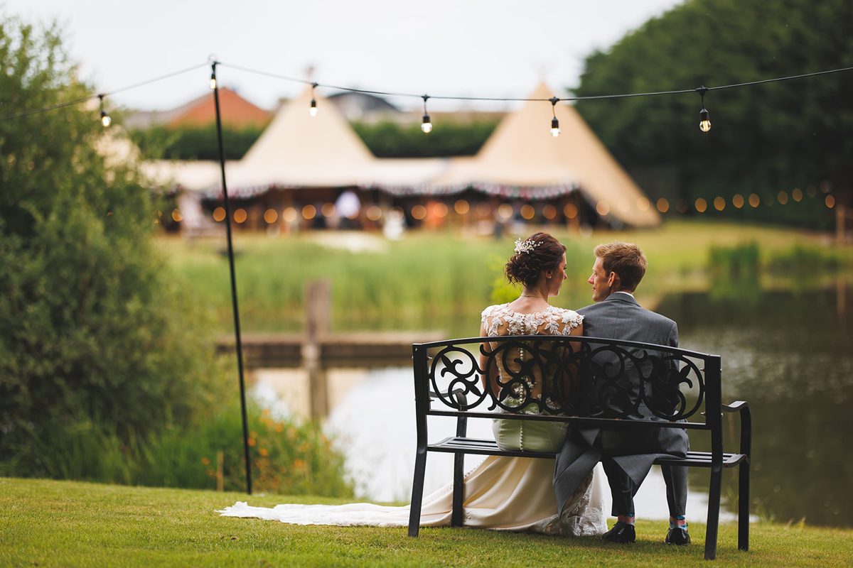 A bride and groom sitting together on a bench by the lake under festoon lights, looking towards two linked Java tipis