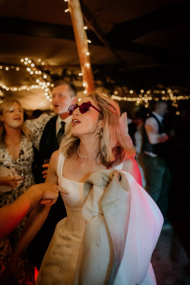 A bride in heart shaped sunglasses dancing with her guests under fairy lights inside a Java tipi