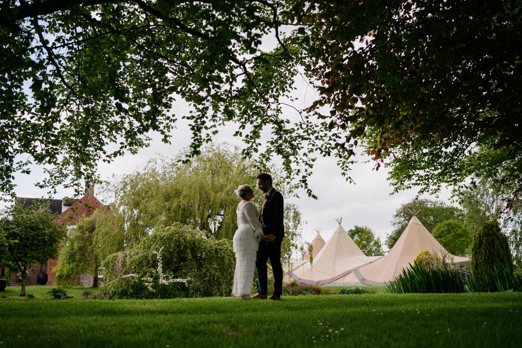 A couple under the trees in the gardens at Winsley Park Farm, with Java tipis and the red brick farmhouse beyond