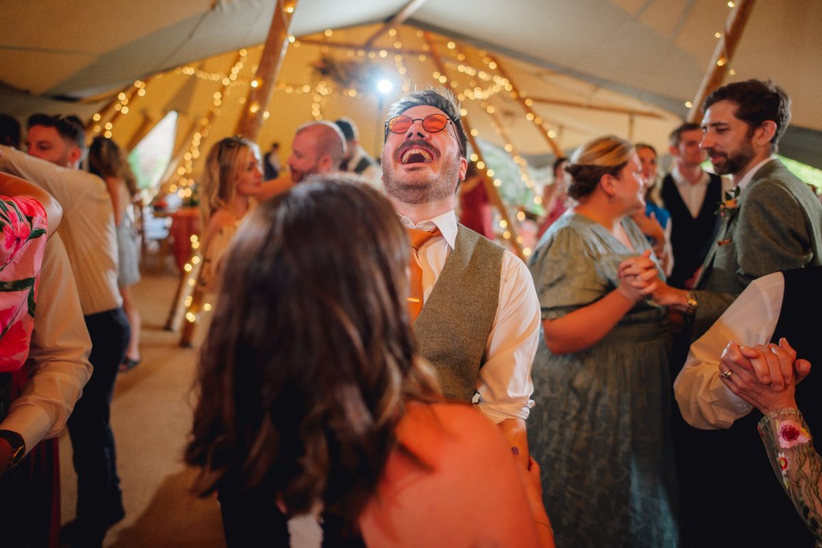 A guest in orange tinted glasses laughing on the dancefloor among friends under fairy lit poles inside a Java tipi