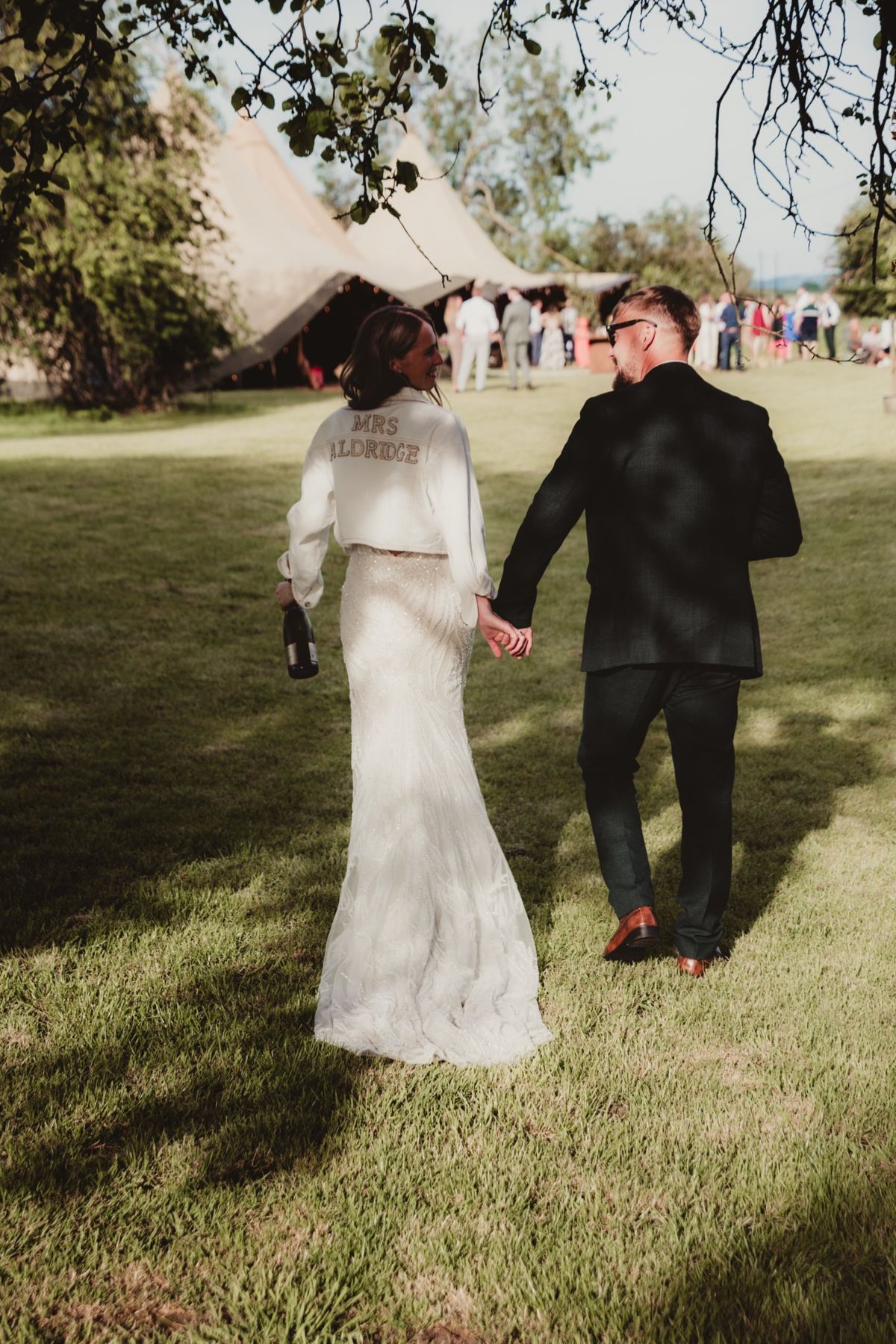 Bride in a Mrs Aldridge jacket walking hand in hand with her groom towards the tipis