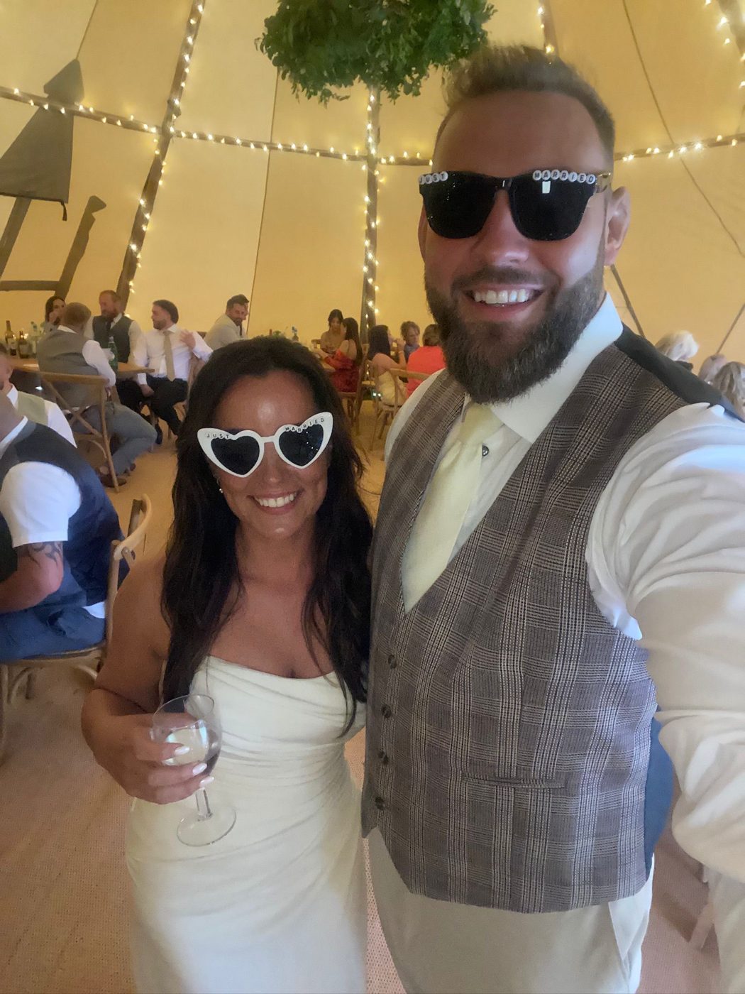 Emma and Jonny in Just Married sunglasses inside their fairy lit tipi, with guests at the tables behind