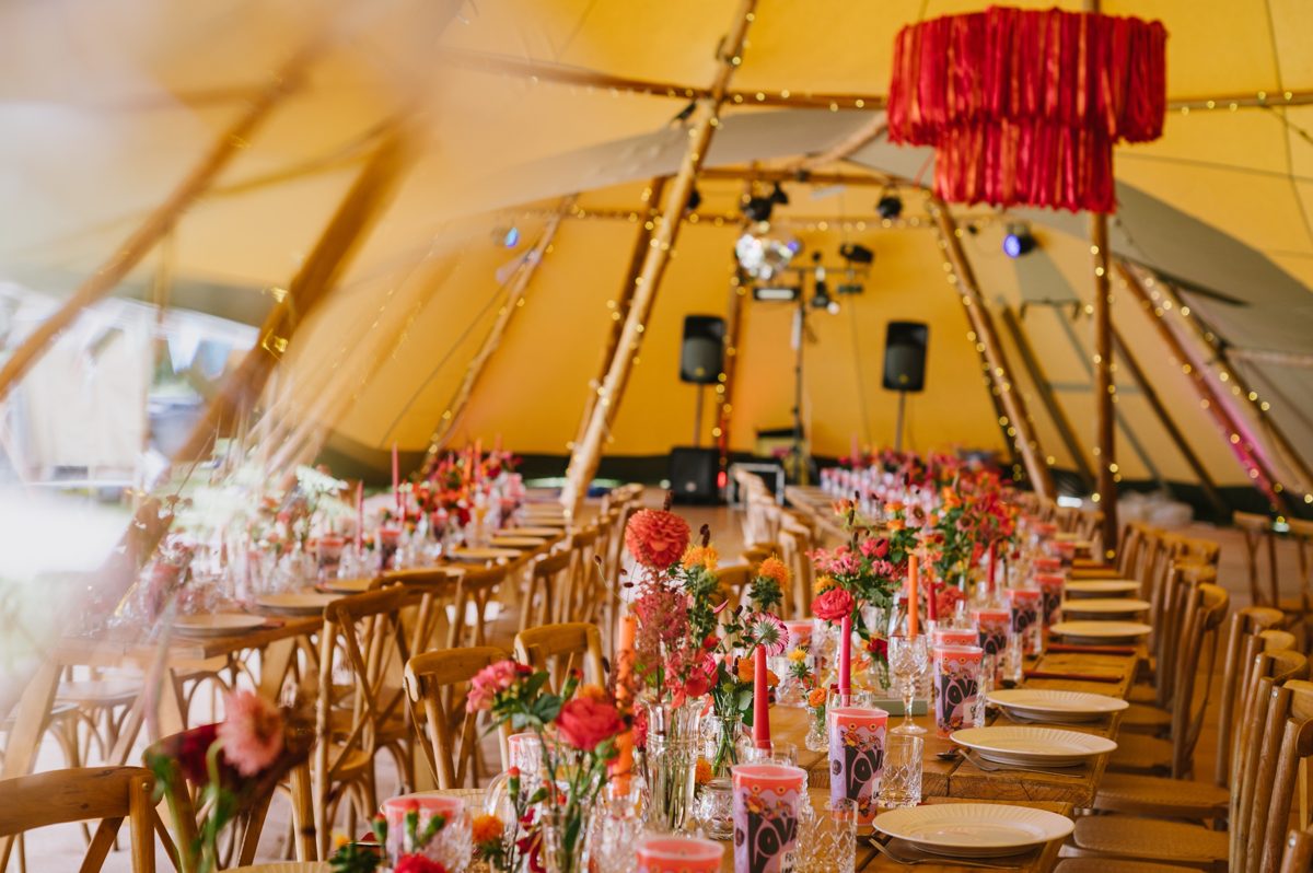Festival vibes. Long tables with bright orange and pink flowers, taper candles and a red ribbon chandelier inside linked Java tipis, with the band set up beyond