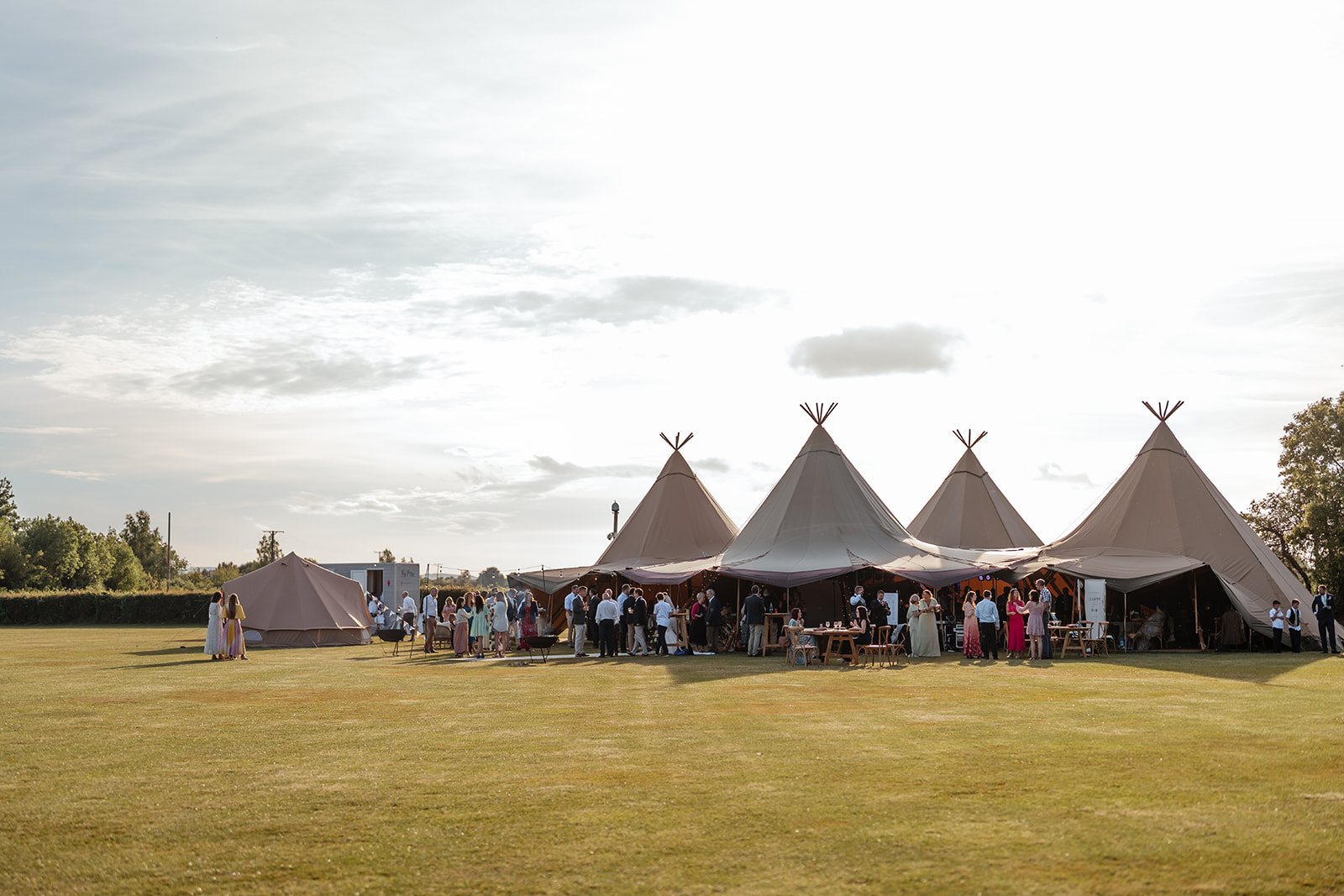 Four linked Java tipis on a lawn with guests and a bell tent
