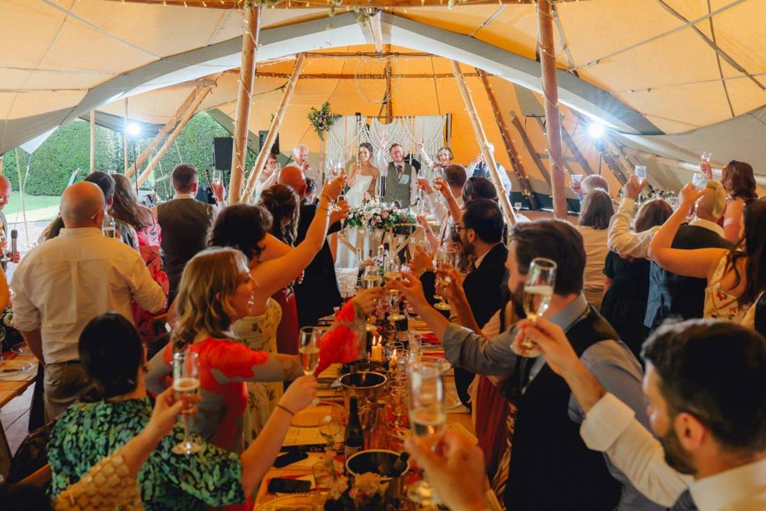Guests at long tables raising a toast to the bride and groom at their top table, with a macrame backdrop, inside linked Java tipis