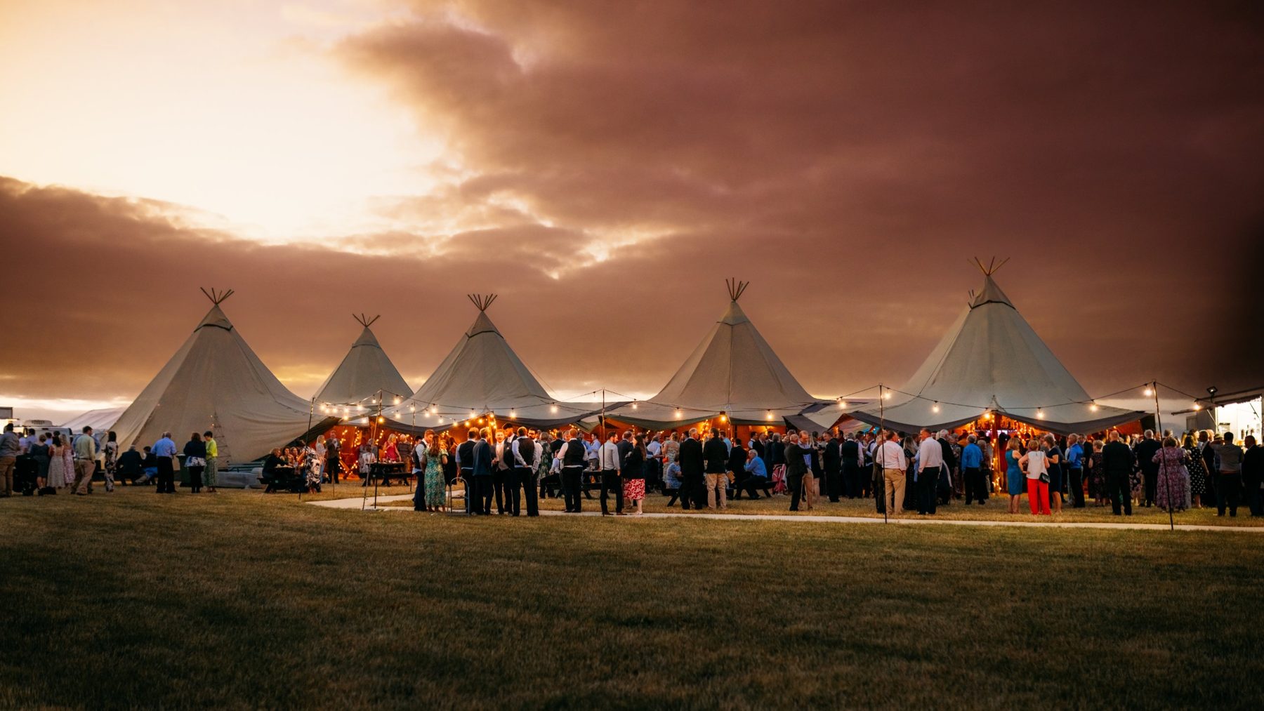 Holly and Tom's guests outside five linked tipis under festoon lights at dusk