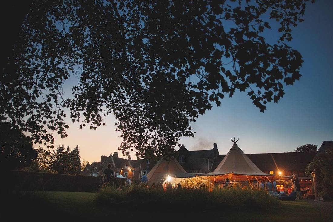 Java tipis glowing with fairy lights at dusk beneath a silhouetted tree, in the gardens of a historic manor house