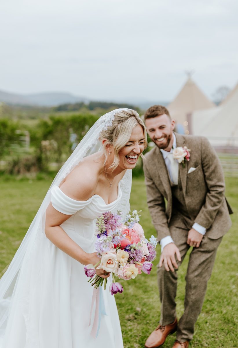Laughing lasts a lifetime. A bride with a bright bouquet and her groom laughing together on the lawn, with Java tipis behind them