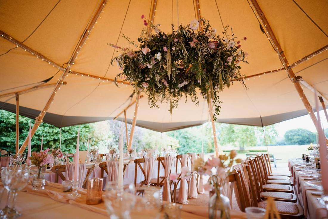 Long tables styled in blush linen with crossback chairs, taper candles and a hanging floral installation inside a fairy lit Java tipi