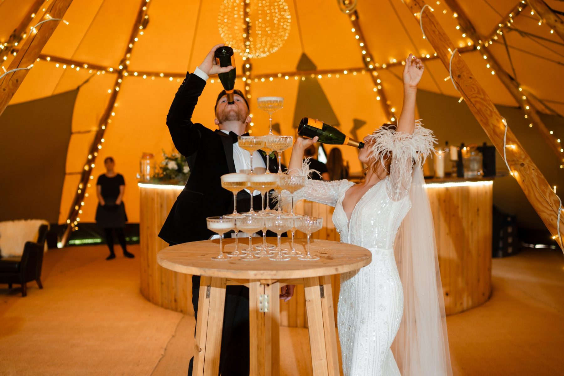 Martha and Daniel drinking champagne from the bottle beside a champagne tower inside their fairy lit tipi