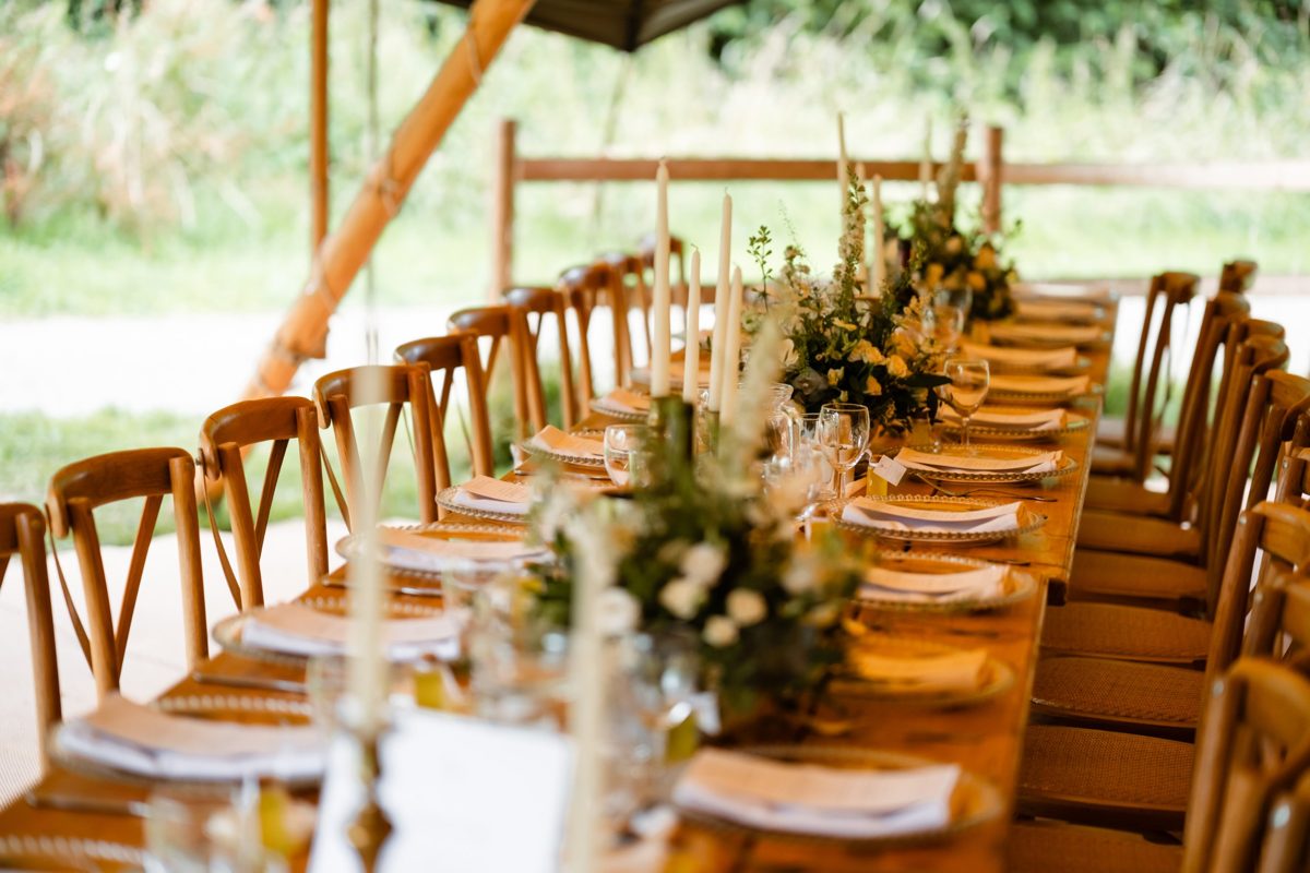 Simple styling. A long wooden table with crossback chairs, white taper candles, glass charger plates and soft green and white flowers inside an open sided Java tipi