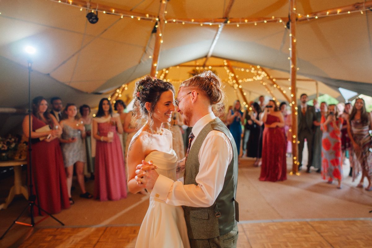The bride and groom smiling at each other during their first dance, with guests watching under fairy lit poles inside a Java tipi