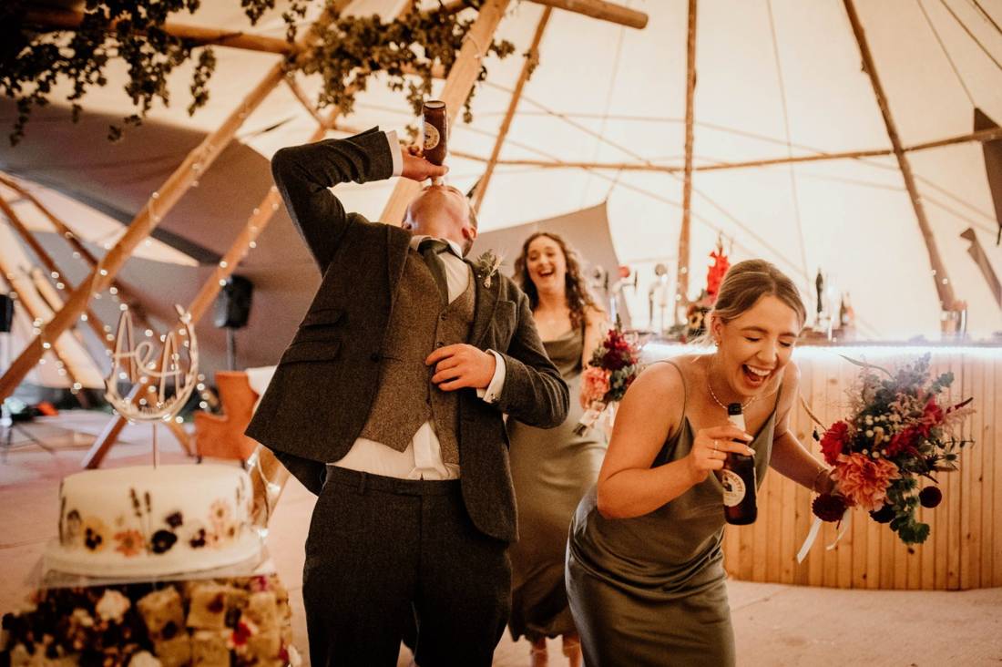 The groom downing a drink as laughing bridesmaids with bouquets dance past the wedding cake inside a fairy lit Java tipi, with our round bar behind