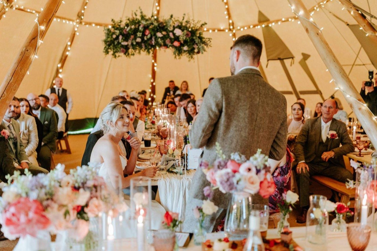 The groom giving his speech to the bride and guests at long tables under a hanging floral installation inside a fairy lit Java tipi